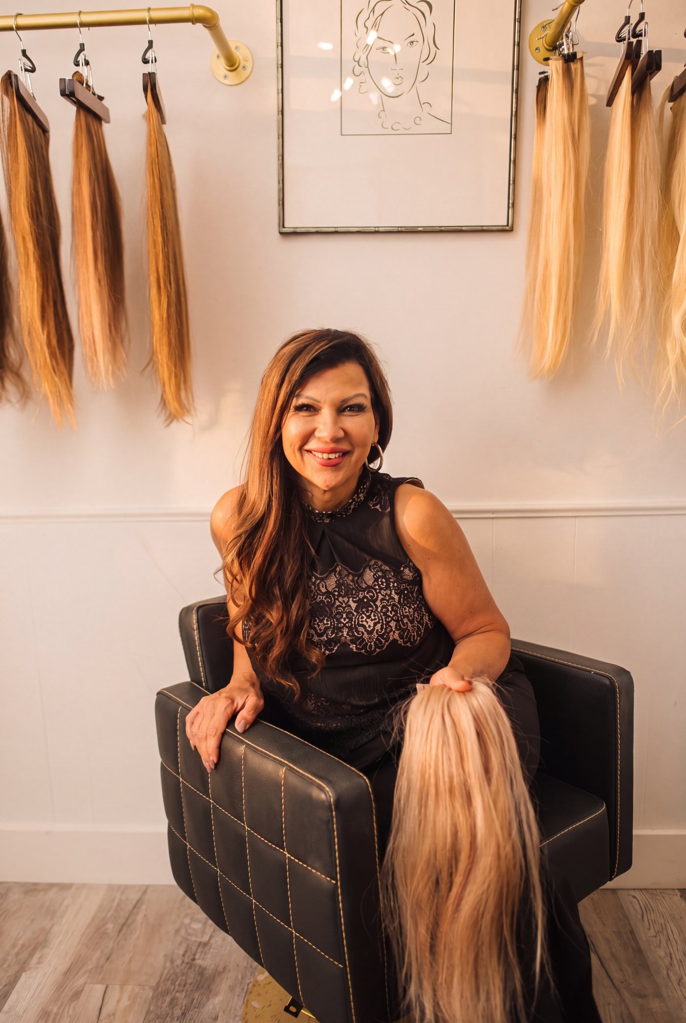Brittany Molina seated with a hand-tied blonde weft, her extension collection on the wall